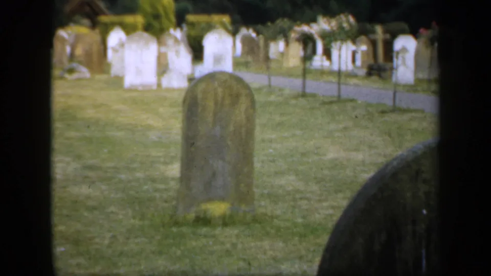 1949 England home-movie frame showing a quiet churchyard with weathered headstones, grass, and a path beyond them.