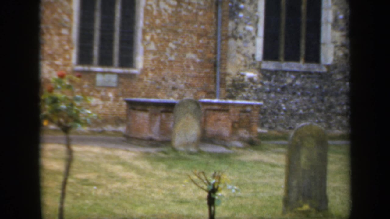 1949 England 8mm frame showing gravestones on a grassy churchyard beside brick and flint masonry with leaded windows.