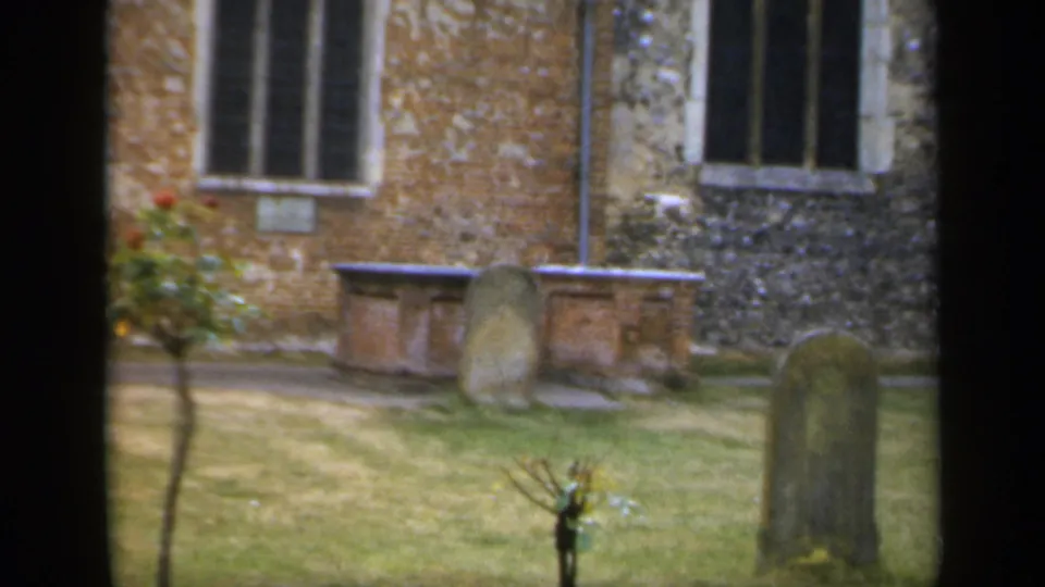 1949 England 8mm frame showing gravestones on a grassy churchyard beside brick and flint masonry with leaded windows.