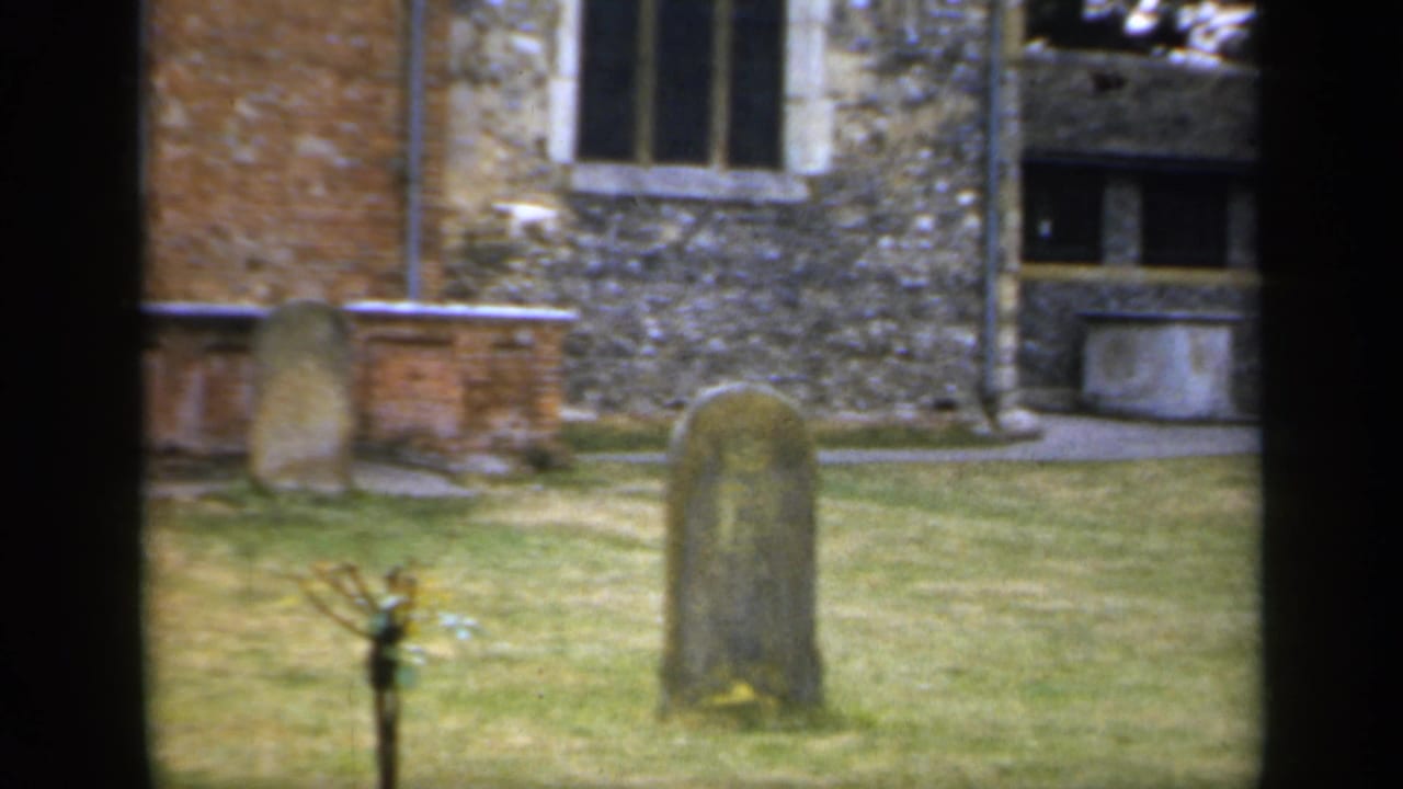 1949 England 8mm frame with old gravestones, a small tree, grass, and mixed brick and flint church masonry in view.
