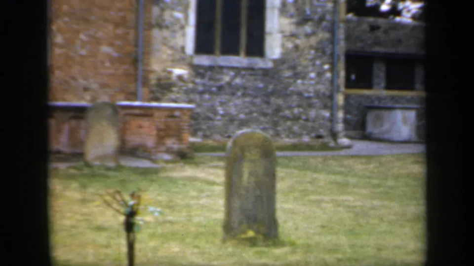 1949 England 8mm frame with old gravestones, a small tree, grass, and mixed brick and flint church masonry in view.