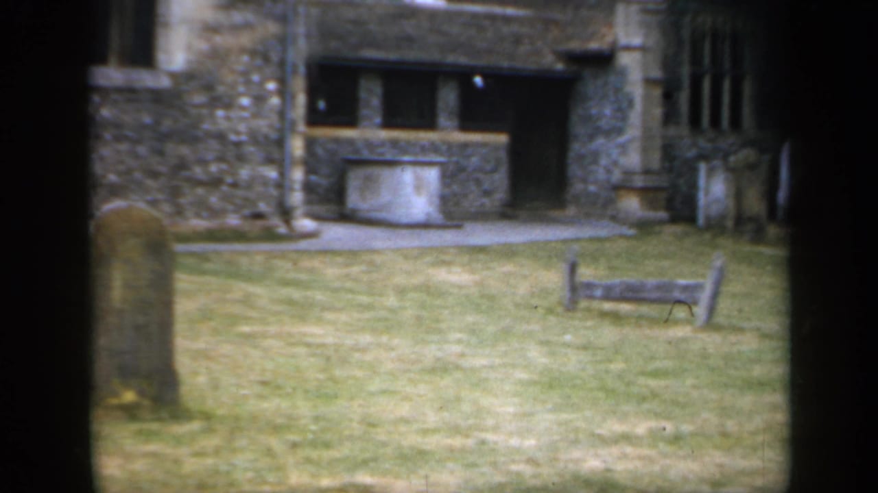 1949 England 8mm frame showing gravestones, lawn, a stone feature near the church wall, and dark frame edges from the film scan.