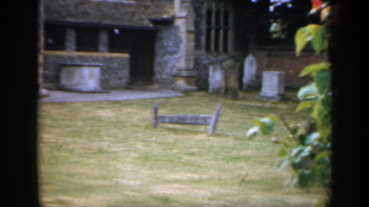 1949 England home-movie frame showing a stone building, lawn, wooden bench, and gravestones in quiet grounds.