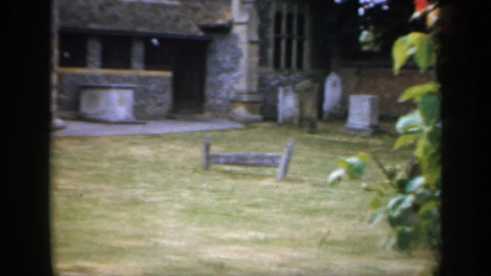 1949 England home-movie frame showing a stone building, lawn, wooden bench, and gravestones in quiet grounds.
