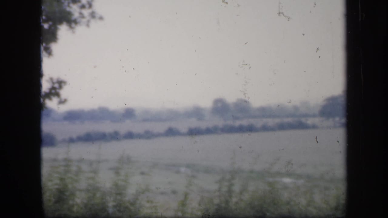 1949 England 8mm frame of a pale rural field scene viewed through glass, with hedgerows and distant trees on the horizon.