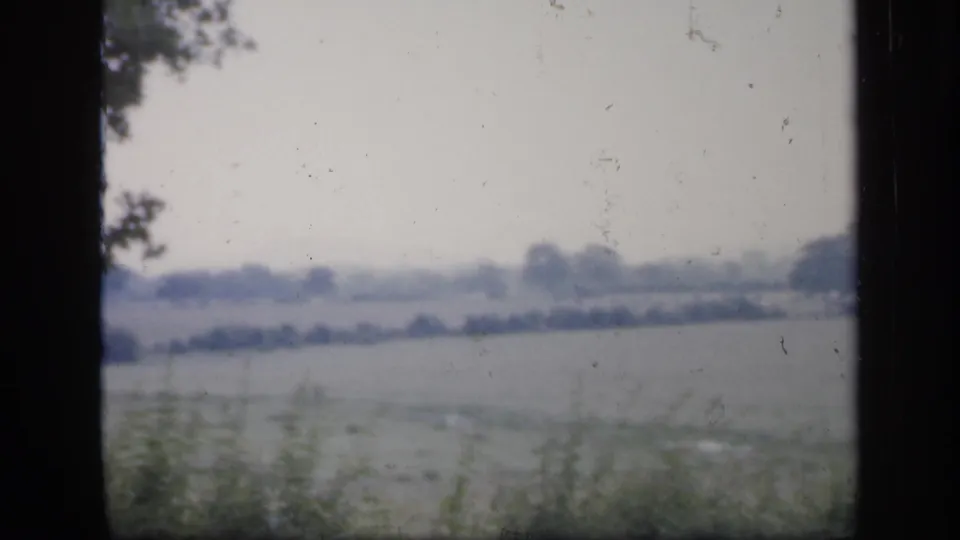 1949 England 8mm frame of a pale rural field scene viewed through glass, with hedgerows and distant trees on the horizon.