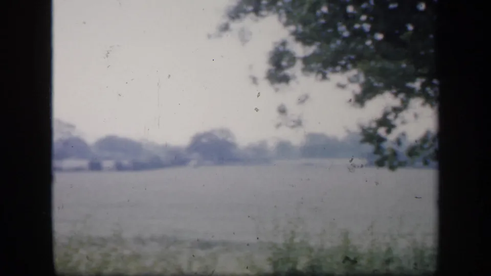 1949 England home-movie frame with soft exposure, showing a roadside edge, distant field, trees, and dark film-frame borders.