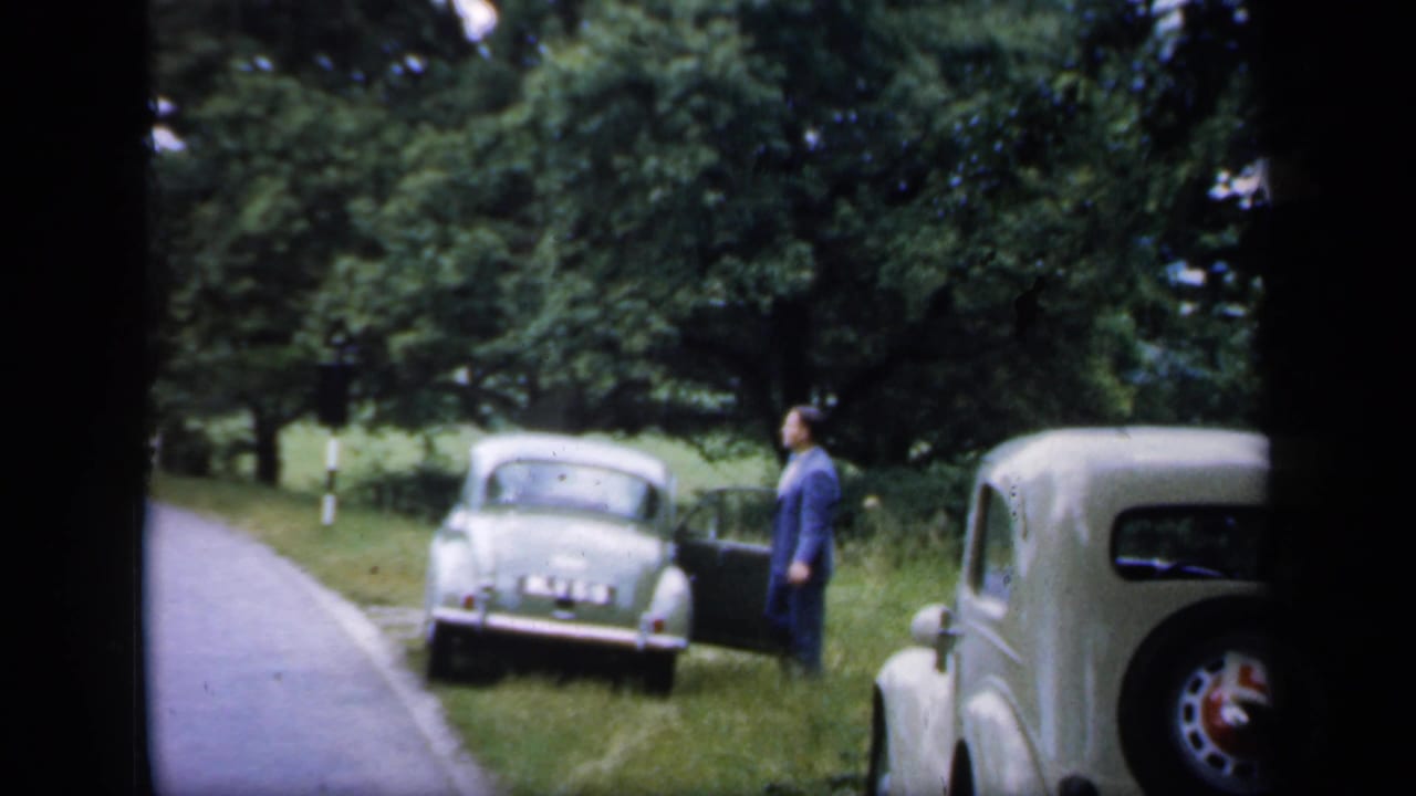 1949 England rural roadside view with a suited man near a light green vintage car and a cream car at the right edge.