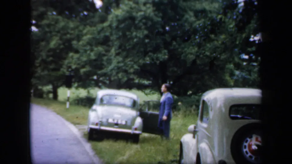 1949 England rural roadside view with a suited man near a light green vintage car and a cream car at the right edge.