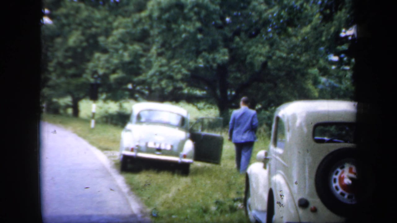 1949 England home-movie frame with a man by an open vintage car door and a cream car parked in the foreground.
