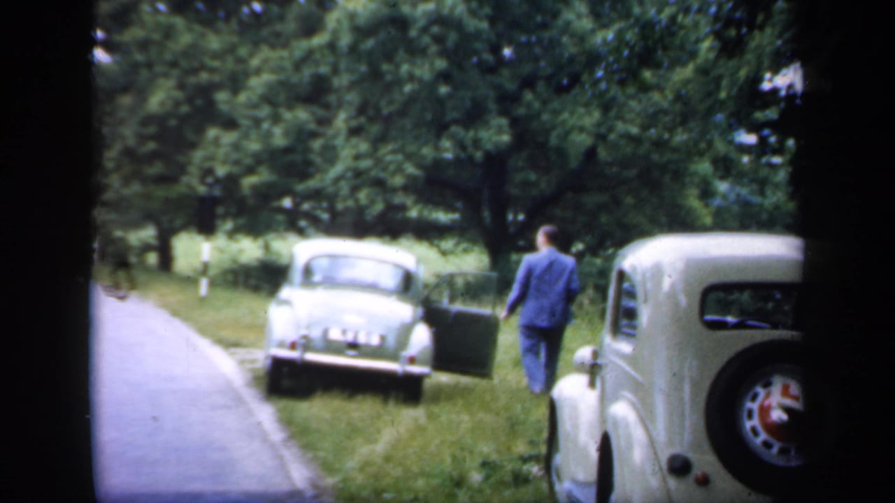 Man in a blue suit stands near a light car beside a rural road, with another classic car in the foreground.