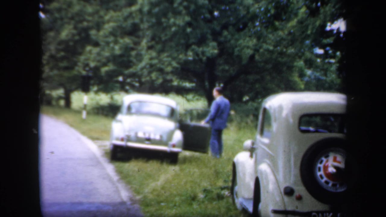 A 1949 England roadside scene shows a man near a parked car, with another classic car close to the camera and trees beyond.