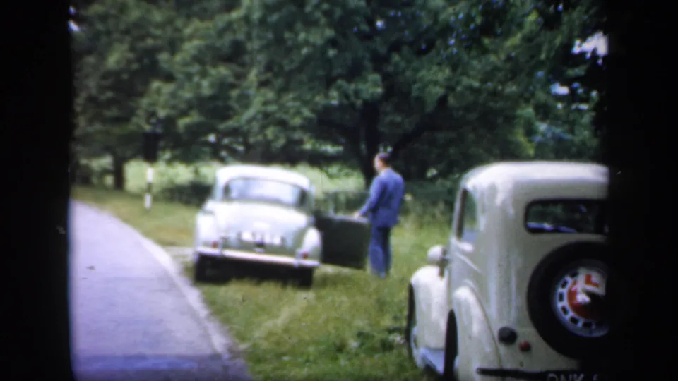 A 1949 England roadside scene shows a man near a parked car, with another classic car close to the camera and trees beyond.
