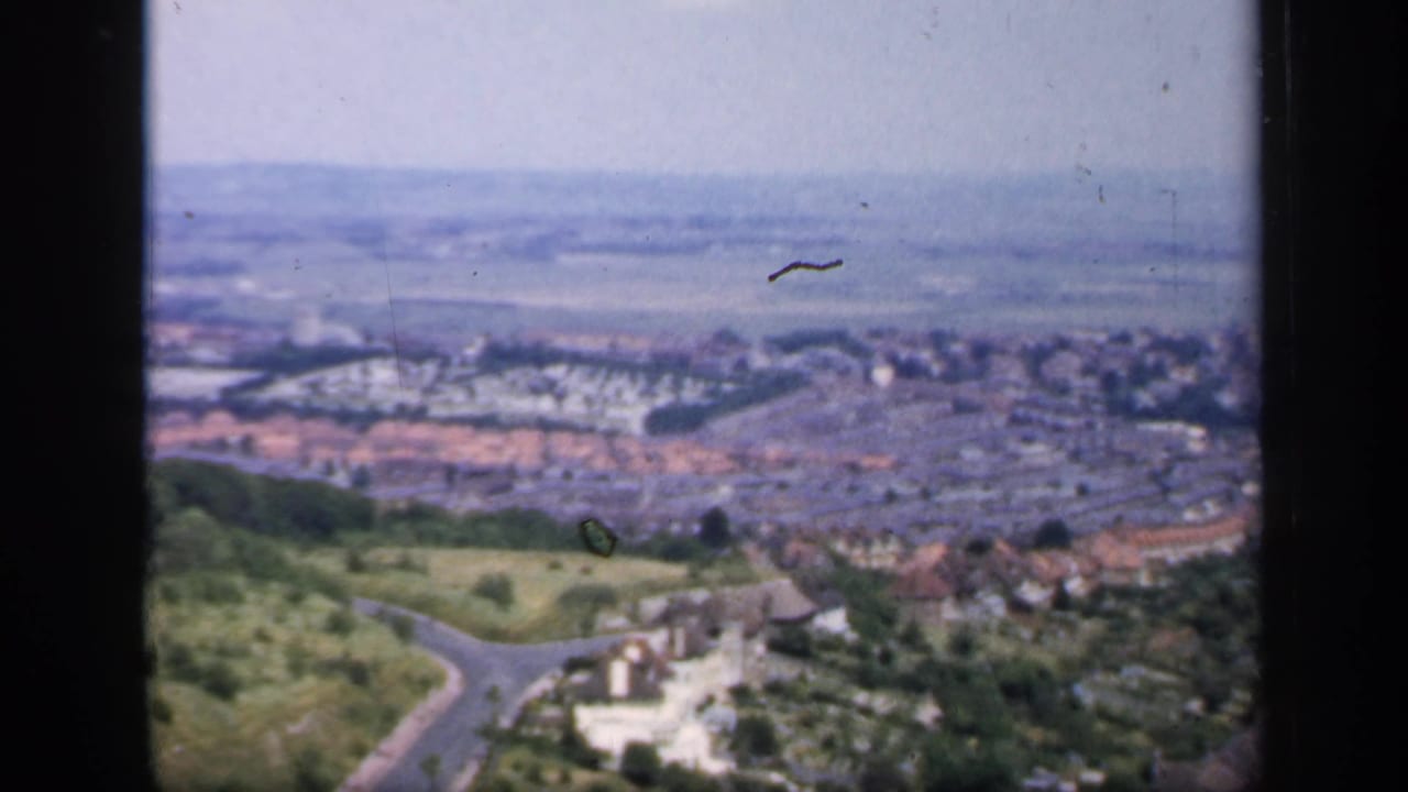 1949 England hilltop pan showing a town edge, green slopes, winding roads, red-roofed houses, and fields beyond.