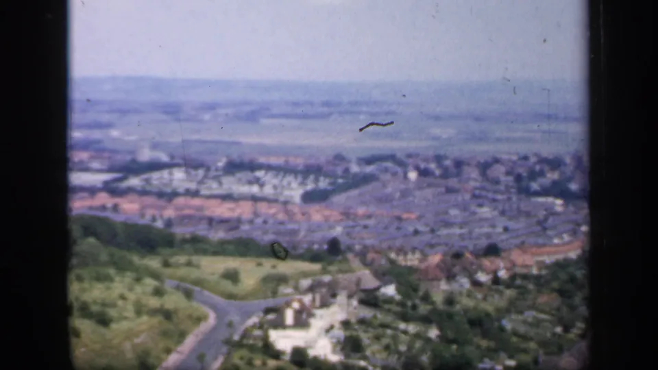 1949 England hilltop pan showing a town edge, green slopes, winding roads, red-roofed houses, and fields beyond.