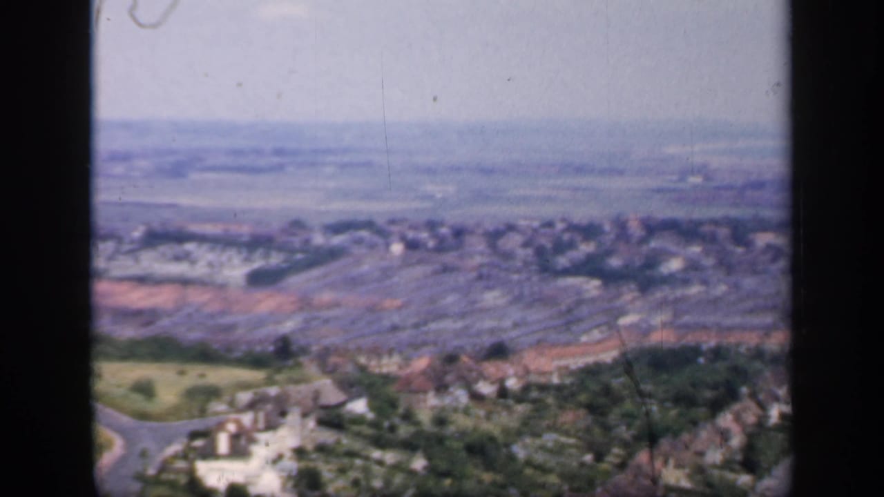 1949 8mm frame of an English town seen from above, with rooftops, hillside vegetation, a road, and fields in soft focus.