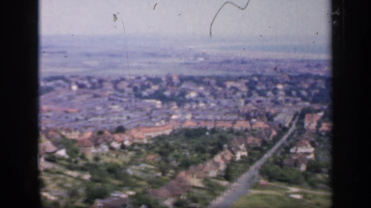 1949 England home-movie frame looking from a hill across clustered houses, a road, trees, and open fields in the distance.