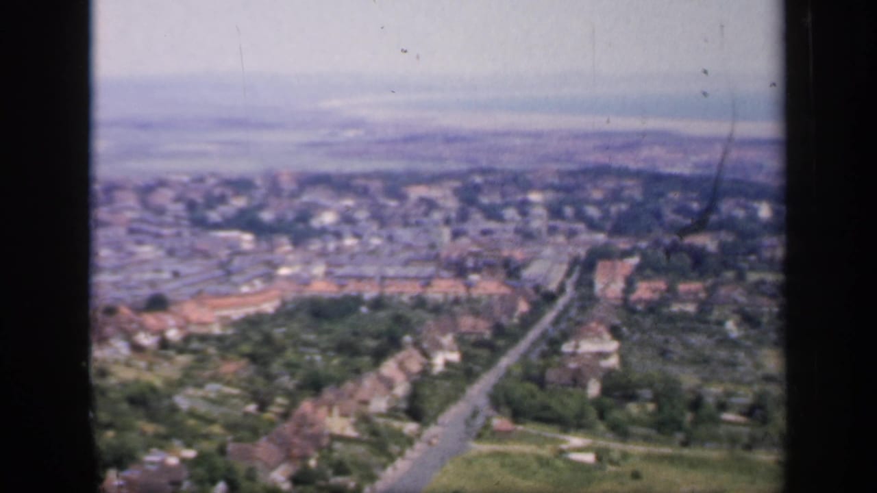 Soft aerial view over houses, roads, trees, and fields under a pale sky.