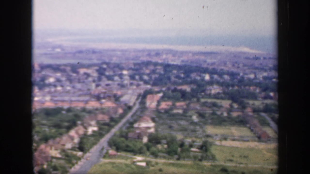 1949 England 8mm frame looking across suburban houses, a main road, trees, fields, and a hazy distant townscape.