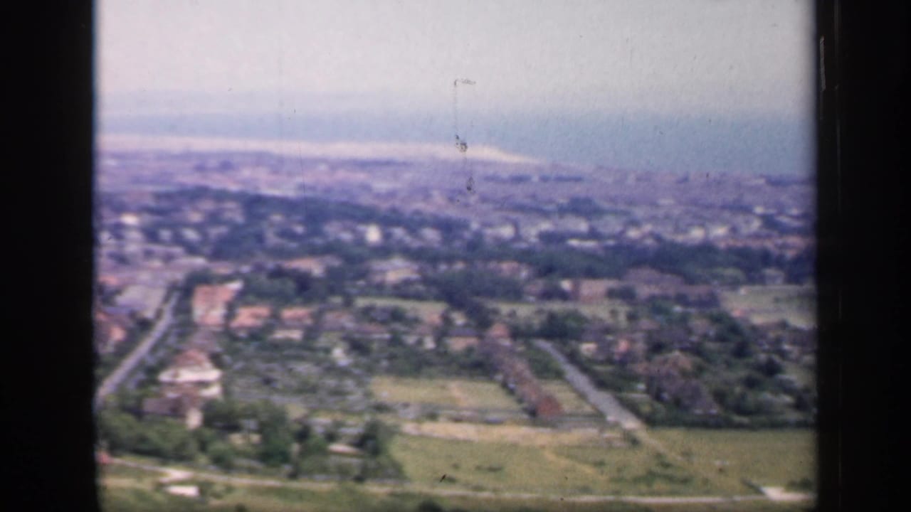 1949 England aerial home-movie frame showing fields in the foreground, roads and houses below, and a hazy town beyond.