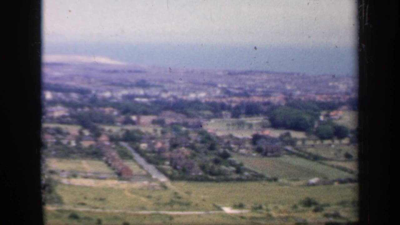 1949 England 8mm aerial frame with open farmland in front, scattered homes and roads below, and a hazy town in the distance.