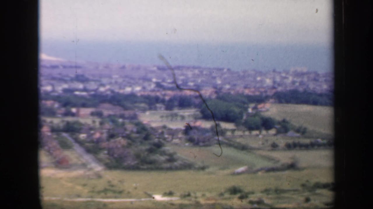 Elevated view over green fields toward a hazy coastal town and sea horizon.