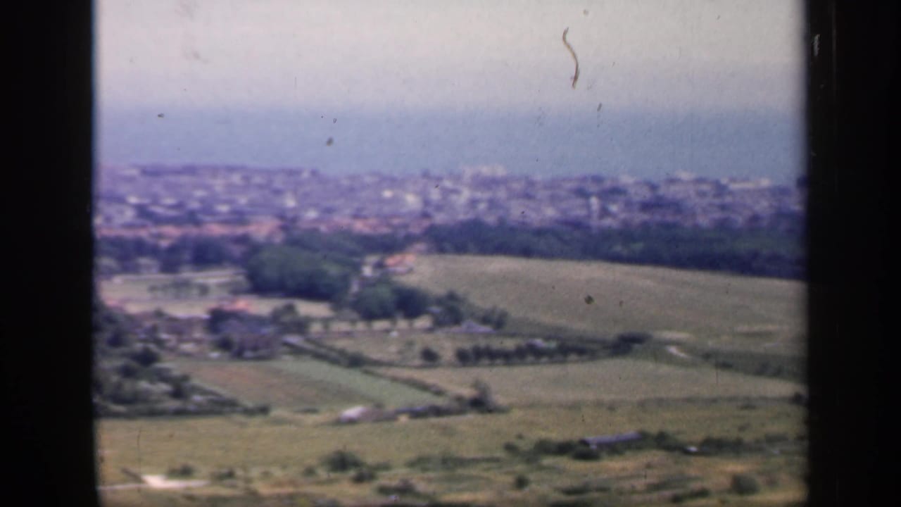1949 England home-movie view of rolling farmland, wooded areas, and a distant coastal town under a pale sky.