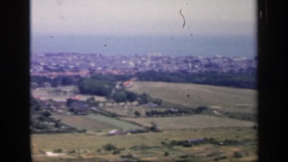 1949 England home-movie view of rolling farmland, wooded areas, and a distant coastal town under a pale sky.