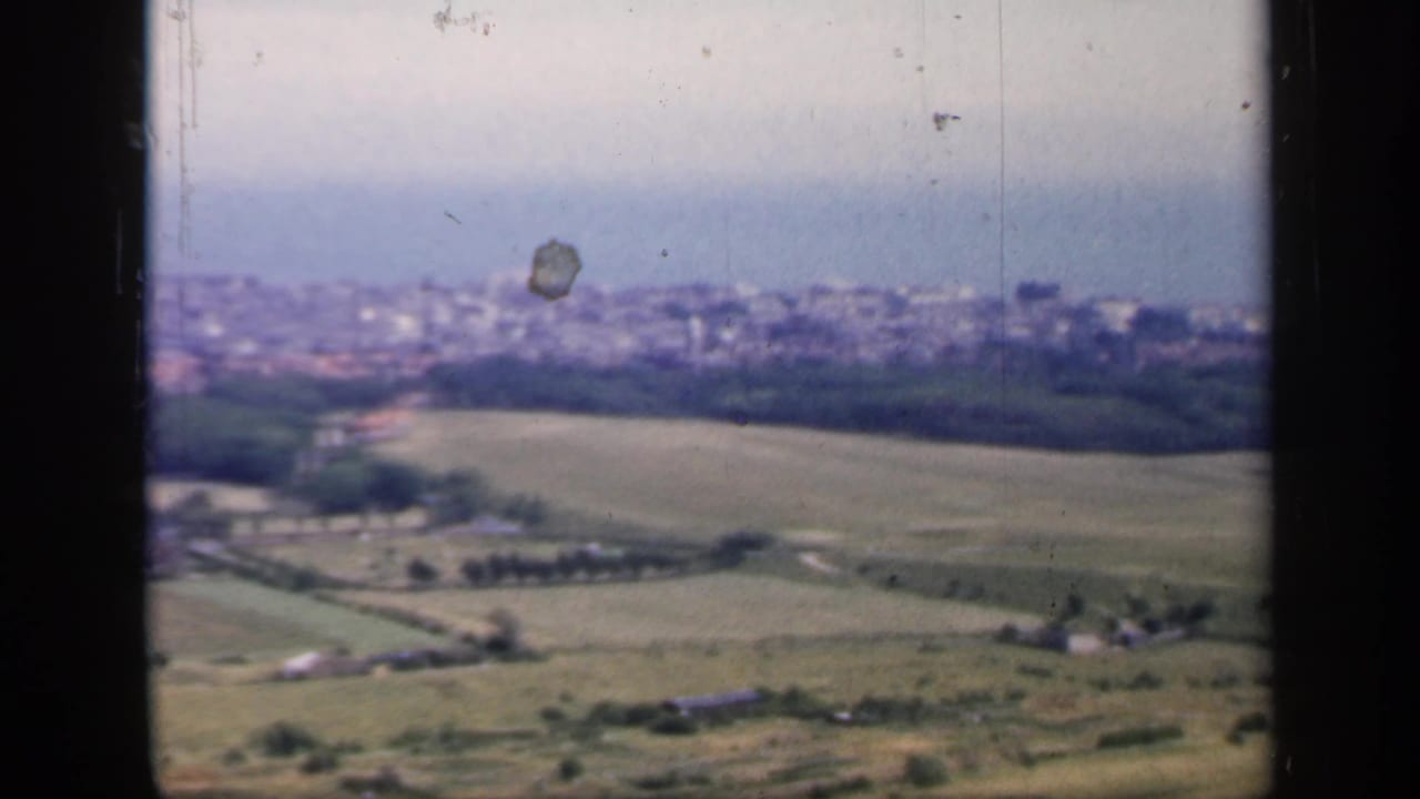 A soft 8mm frame from 1949 England looking over open fields and farm structures toward a hazy coastal settlement.