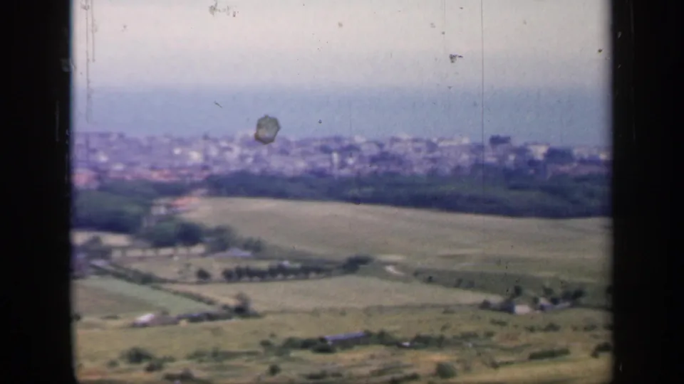 A soft 8mm frame from 1949 England looking over open fields and farm structures toward a hazy coastal settlement.