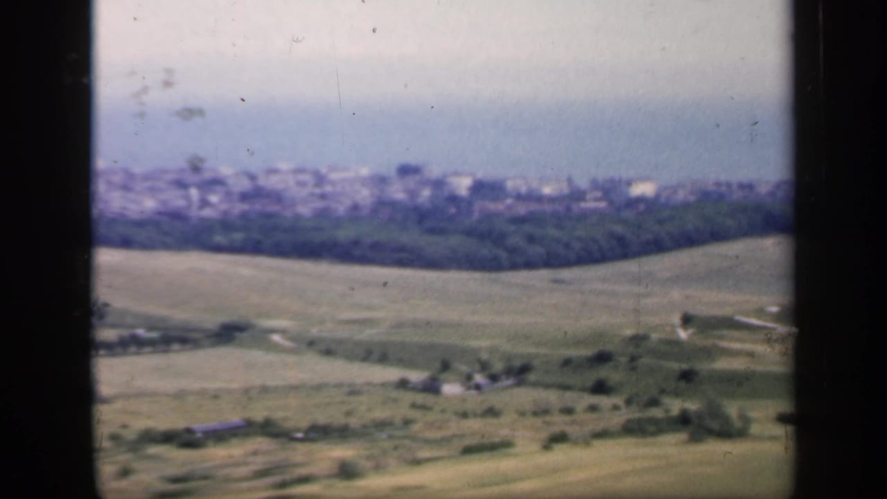 End frame of the 1949 England view, with sloping pasture, trees, scattered buildings, and a hazy town near the sea.