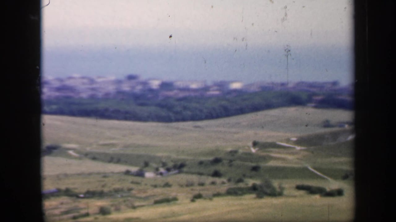 Rolling green hills with distant town buildings under a pale hazy sky.
