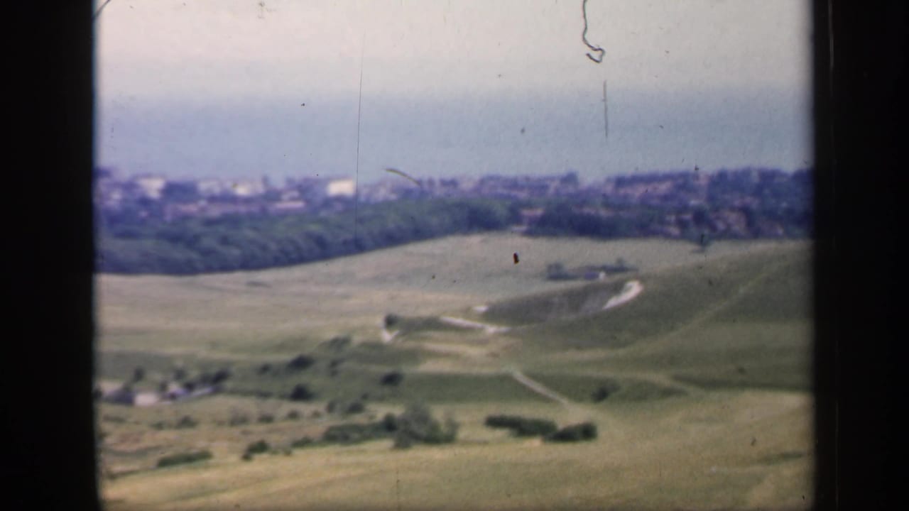 1949 England countryside scene with sloped green fields, winding paths, tree belts, and distant town buildings.