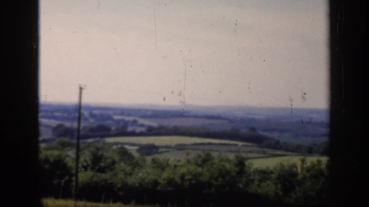 Rolling green farmland and hedgerows seen through a dark window frame.
