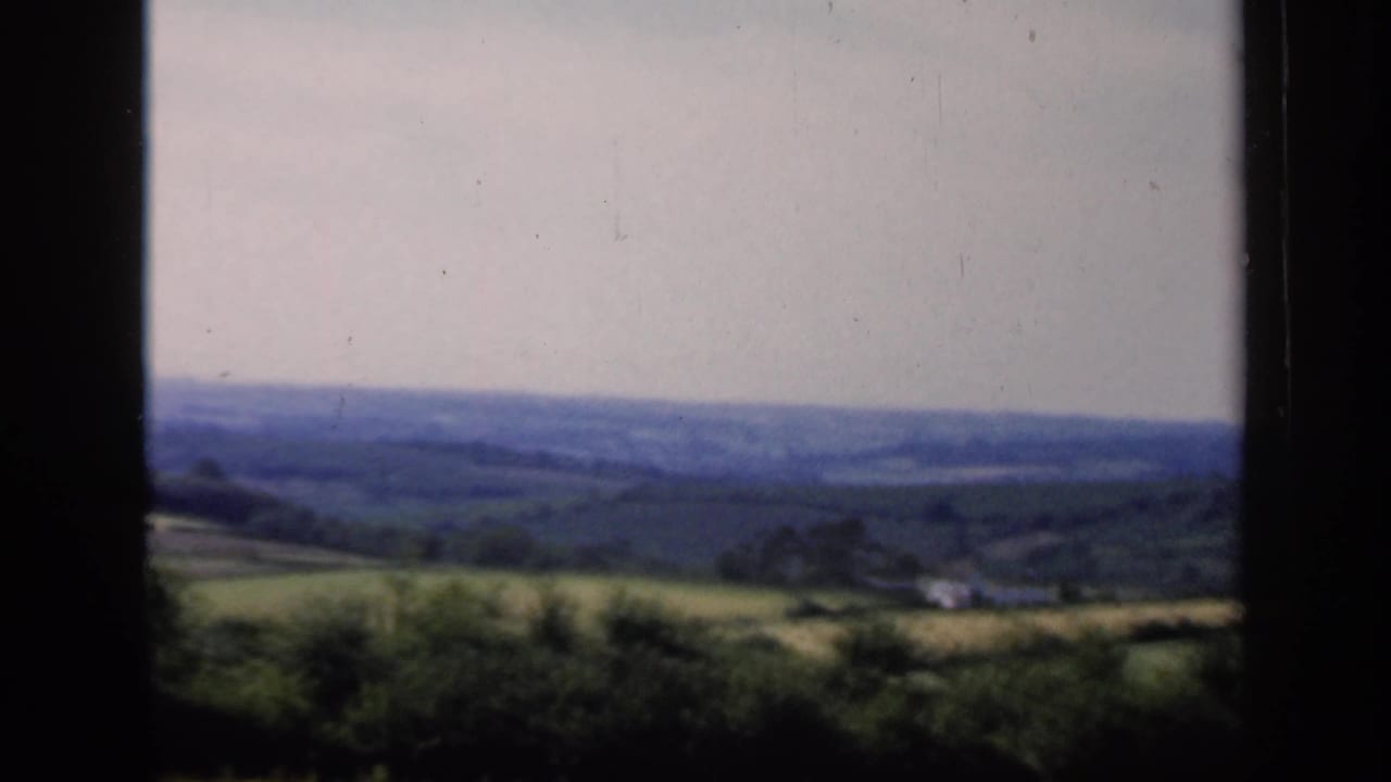 1949 England countryside frame showing soft-focus hills, fields, hedgerows, and a small pale structure in the distance.