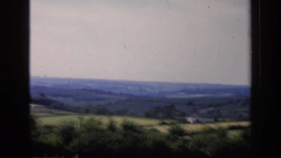 1949 England countryside frame showing soft-focus hills, fields, hedgerows, and a small pale structure in the distance.