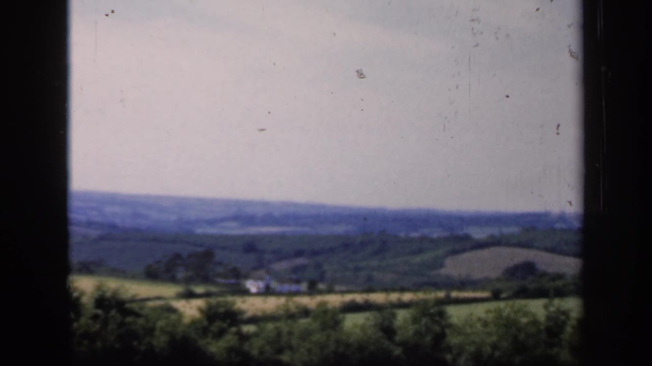 Late-1940s England home-movie view of farmland, hedgerows, distant hills, and small rural buildings seen from a high vantage point.