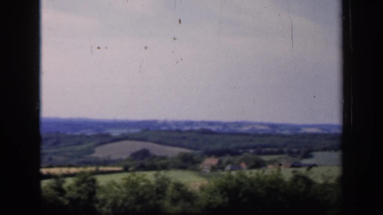 Countryside fields, trees, and low blue hills behind a central dark window bar.