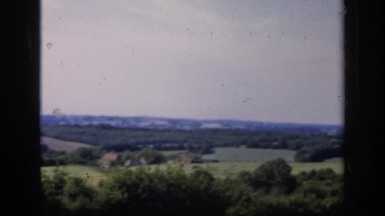 1949 English countryside view with soft 8mm color, green fields, trees, distant hills, and small structures near the horizon.
