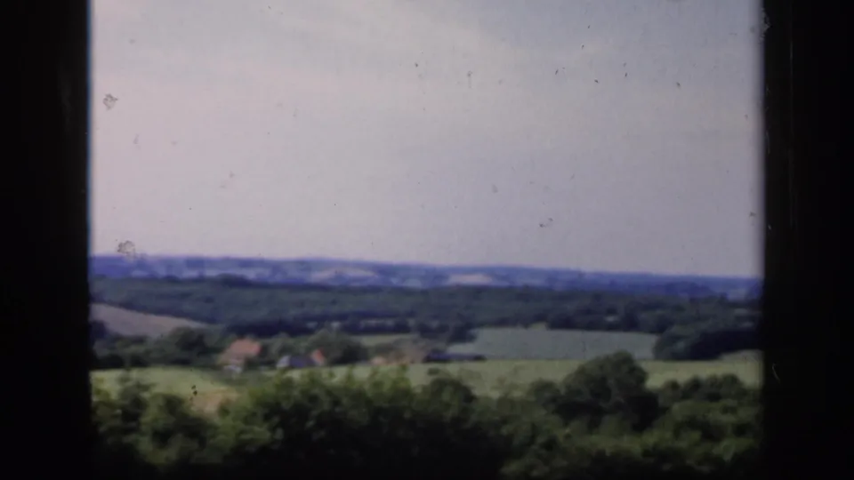 1949 English countryside view with soft 8mm color, green fields, trees, distant hills, and small structures near the horizon.