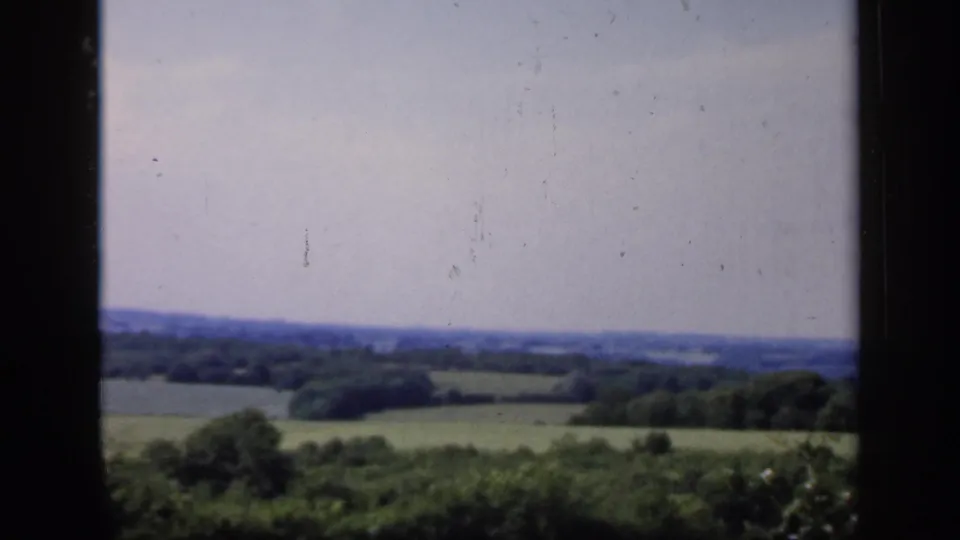 1949 England 8mm frame showing rolling green fields and distant trees viewed through a dark interior window frame.