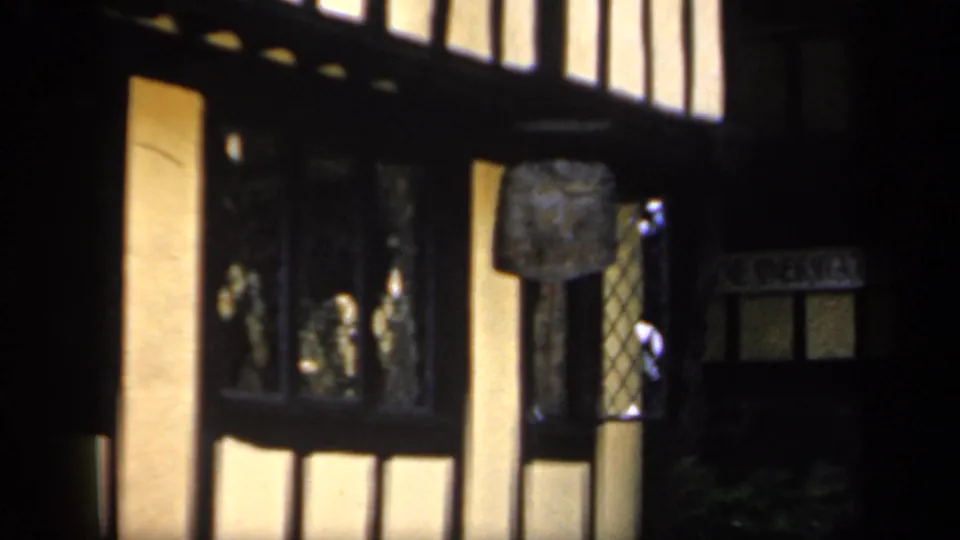 1949 England home-movie frame showing timber framing, pale wall panels, leaded windows, and shadowed signage on a Tudor-style facade.