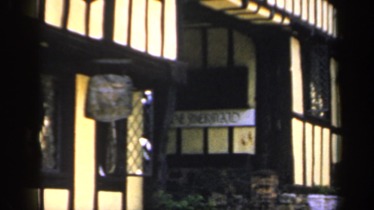 The pan moves across a 1949 England street facade with black beams, yellow plaster, a projecting sign, and partial lettering near the windows.