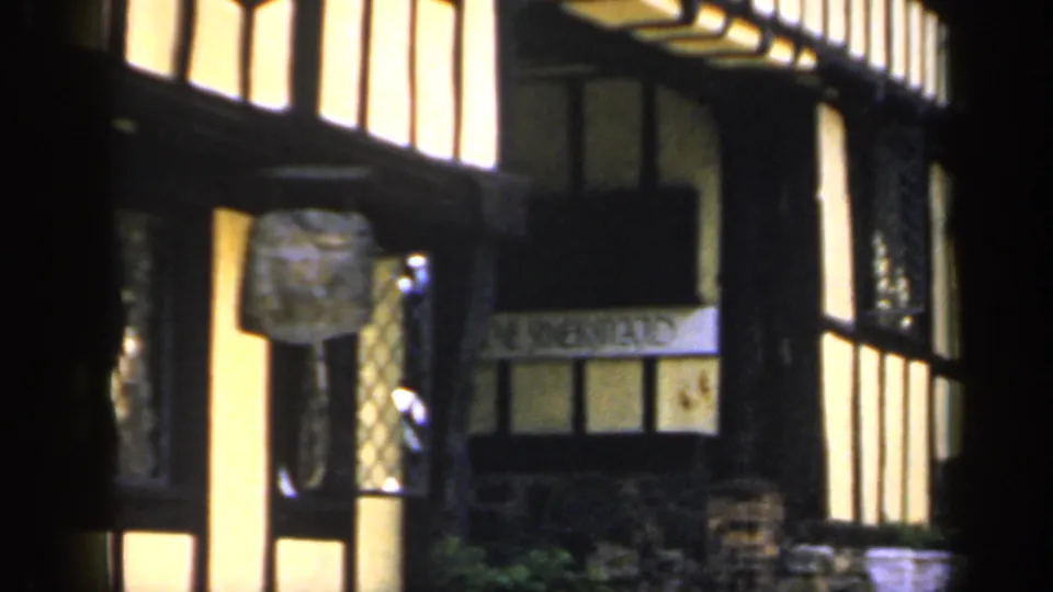 The pan moves across a 1949 England street facade with black beams, yellow plaster, a projecting sign, and partial lettering near the windows.