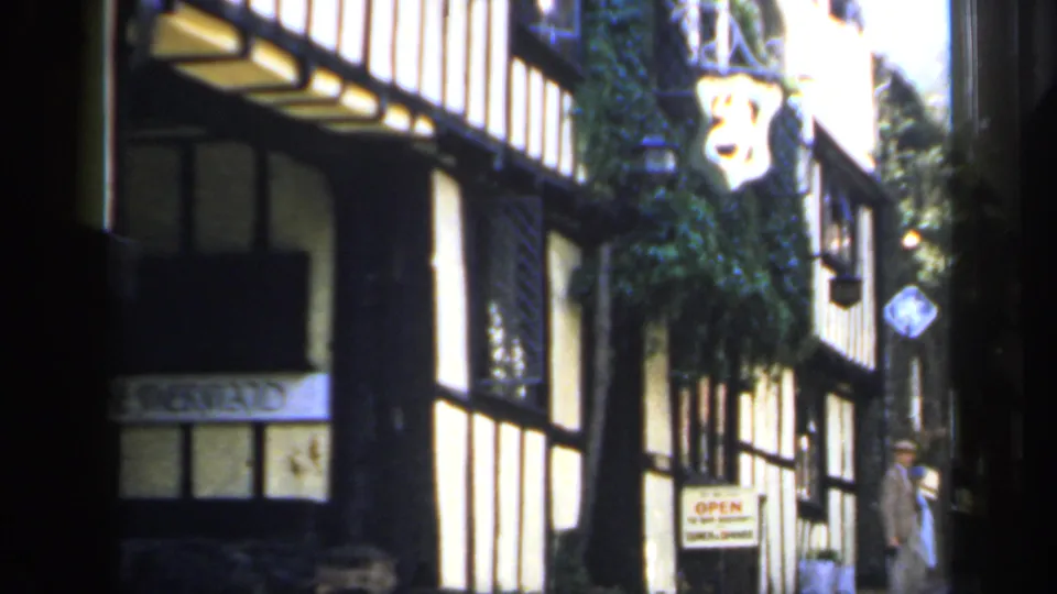 1949 England 8mm frame showing a black-and-white timber-framed shopfront with a hanging sign and shaded windows.