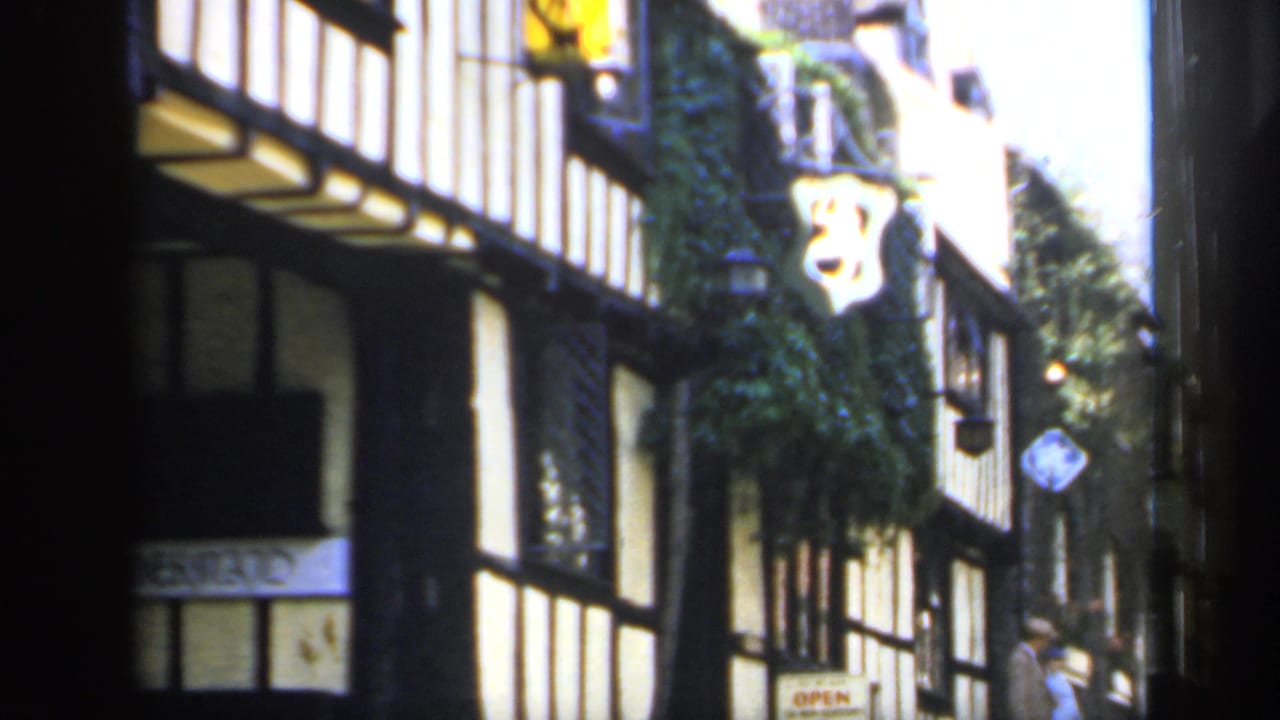 1949 England street view with timber framing, ivy-covered wall, hanging sign, and a passerby in the background.