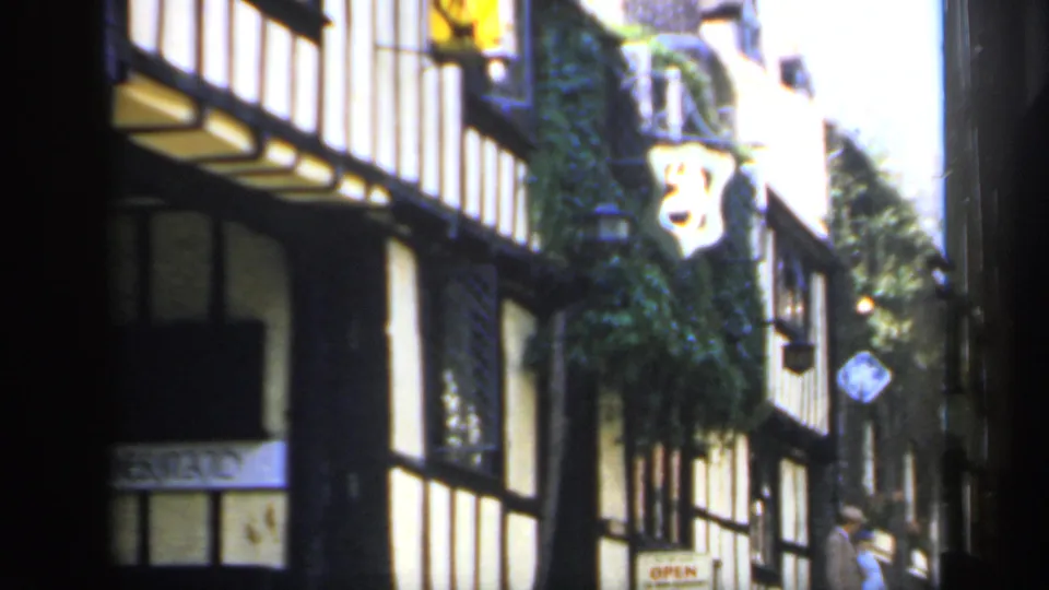 1949 England street view with timber framing, ivy-covered wall, hanging sign, and a passerby in the background.