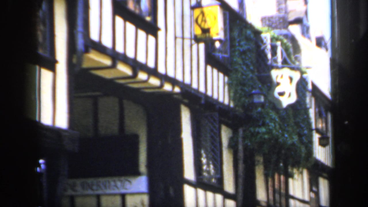 1949 England home-movie frame of a timber-framed shopfront, ivy, hanging sign, and partially visible storefront signage.