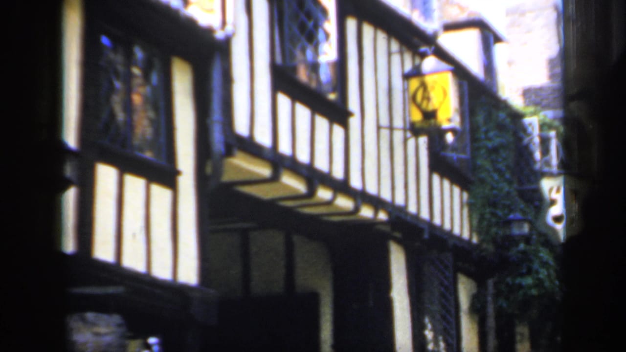 1949 England frame showing an angled timber-framed street facade with projecting windows, ivy, and pub-style hanging signs.