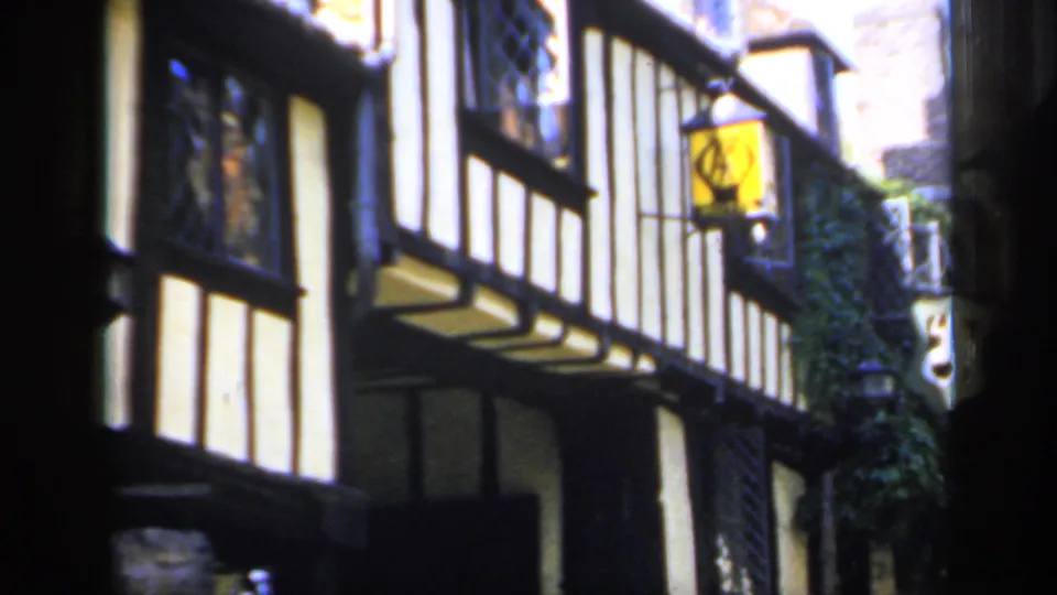 1949 England frame showing an angled timber-framed street facade with projecting windows, ivy, and pub-style hanging signs.